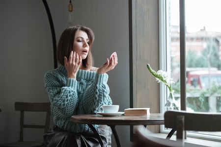 Image of beautiful young lady sitting at the table in cafe and looking at mirror.