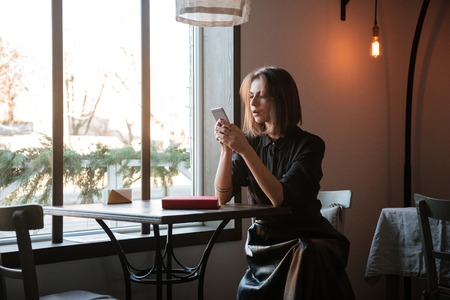 Picture of pretty young lady sitting at the table in cafe and chatting by her phone.