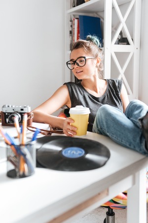 Beautiful young woman in glasses sitting with legs on table and drinking coffeeの写真素材