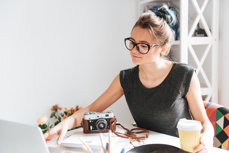 Attractive young woman photogtrapher in glasses drinking coffee and using laptop at the table in the roomの写真素材