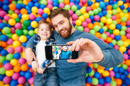 Joyful little boy and dad lying in colorful balls pool and making selfie with mobile phoneの写真素材