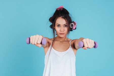Full length portrait of a concentrated young woman with curlers holding weights and looking at camera isolated on a blue backgroundの写真素材