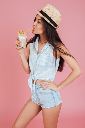 Picture of pretty young woman wearing hat isolated over pink background drinking cocktail.の写真素材
