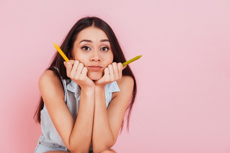 Close up portrait of a confused young woman holding crayons and looking at camera over pink backgroundの写真素材