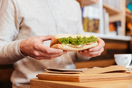 Cropped image of young man dressed in white shirt sitting in cafe while holding sandwich.の写真素材