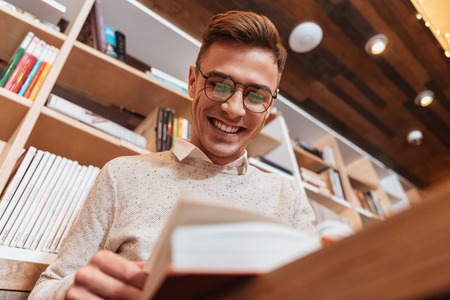 Picture of young happy man wearing glasses dressed in shirt sitting in cafe while reading book. Look...