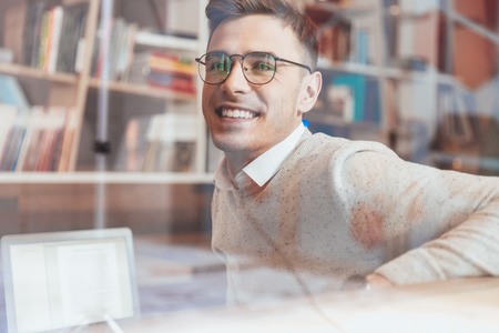 Photo of young cheerful man wearing eyeglasses while sitting in cafe and looking aside.の写真素材
