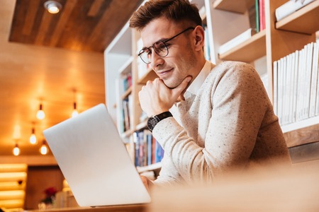 Image of handsome young concentrated man wearing eyeglasses sitting in cafe while using laptop...