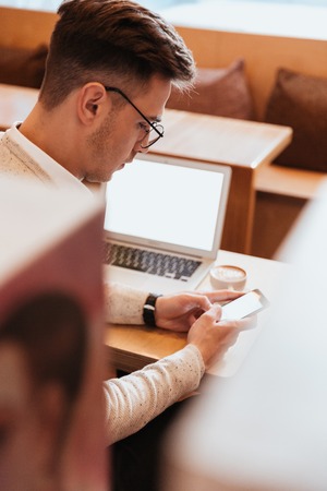 Image of handsome concentrated man sitting in cafe while drinking coffee and using mobile phone. Loking aside.の写真素材