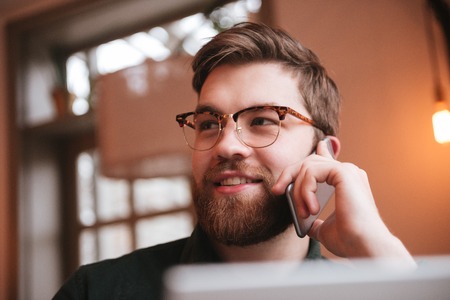 Picture of happy bearded young man wearing glasses sitting in cafe while talking by phone and using laptop.の写真素材