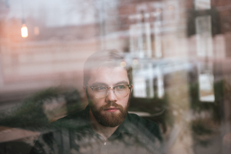 Image of happy bearded young man wearing glasses sitting in cafe while looking at window.の写真素材