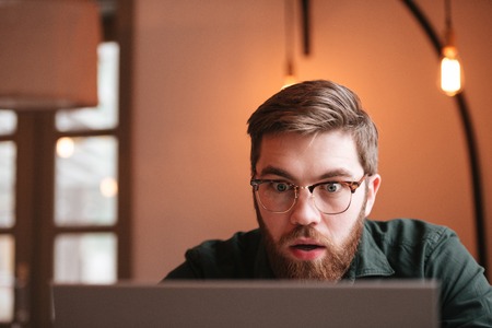 Picture of confused bearded young man wearing glasses sitting in cafe while using laptop computer.の写真素材