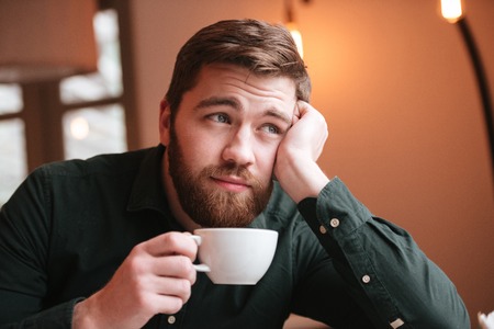 Portrait of sad bearded young man sitting in cafe while drinking coffee.の写真素材