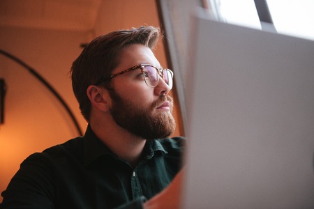 Picture of serious bearded young man wearing glasses sitting in cafe while using laptop.の写真素材