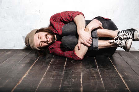 Image of attractive bearded hipster man wearing hat dressed in shirt in a cage lies on floor posing over wall background.の写真素材