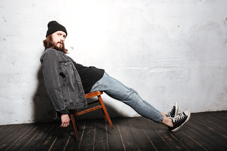Full length portrait of a hipster man in hat sitting on chair isolated over wall backgroundの写真素材