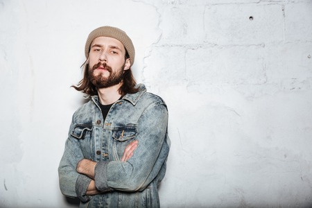 Image of handsome bearded hipster man wearing hat dressed in jeans jacket posing over wall background with arms crossed.の写真素材