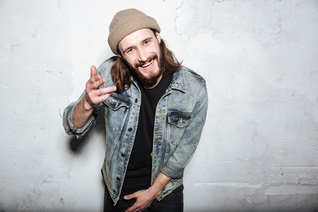 Picture of happy bearded hipster man wearing hat dressed in jeans jacket posing over wall background and gesturing with hands.の写真素材
