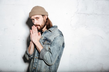 Photo of serious bearded hipster man wearing hat dressed in jeans jacket prays over wall background.の写真素材