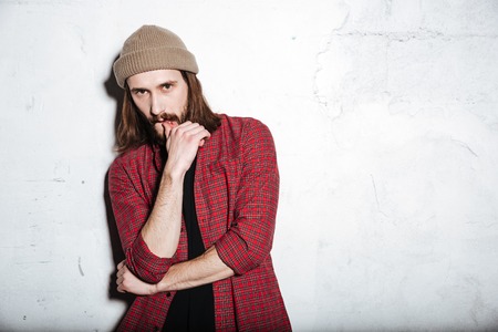 Image of attractive young bearded hipster man wearing hat dressed in shirt in a cage isolated over wall background looking at camera.の写真素材
