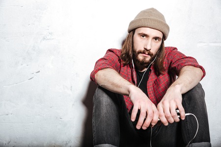 Image of attractive young bearded hipster man wearing hat dressed in shirt in a cage sitting on floor isolated over wall background while listening music with earphones.の写真素材