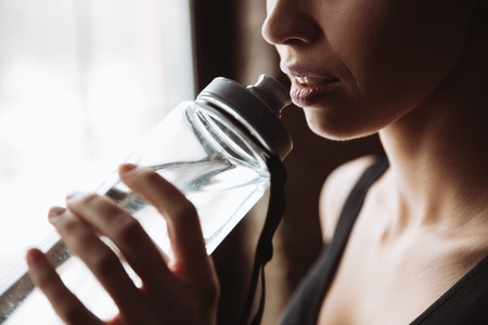 Cropped picture of gorgeous young fitness woman standing in gym near window while drinking water.の写真素材