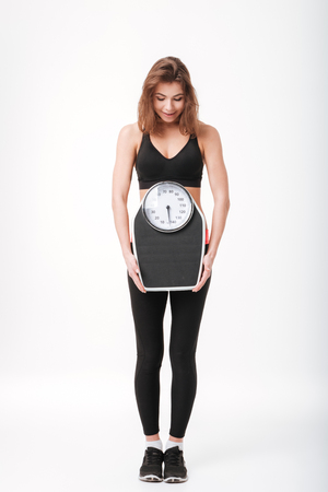 Image of cheerful young fitness lady standing isolated over white background and holding scalesの写真素材