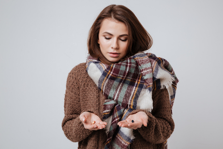 Picture of sick young woman in sweater and scarf choosing medicine pills. Isolated over gray background.の写真素材