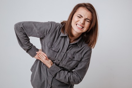 Picture of sick young woman dressed in shirt standing isolated over gray background and touching her body.の写真素材