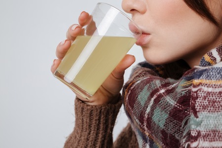 Cropped image of sick young lady in sweater and scarf holding glass with medicine and drinking. Isolated over gray background.の写真素材