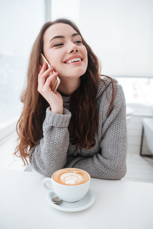 Image of pretty young lady dressed in sweater sitting in cafe at cold winter day and drinking coffee while talking by phone. Look aside.の写真素材