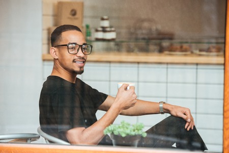 Portrait of a happy afro american man drinking morning coffee at cafe, view through the window