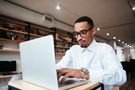 Picture of handsome african businessman wearing glasses sitting at the table typing by laptop computer. Coworking.の写真素材