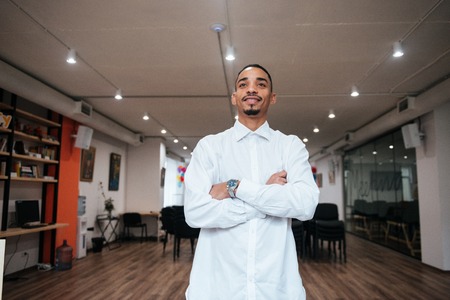 Image of african attractive businessman standing in library with arms crossed. Look aside.の写真素材