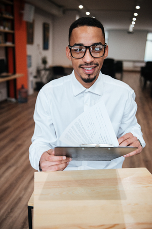 Image of attractive african businessman wearing glasses sitting at the table and looking at notepad. Coworking.の写真素材