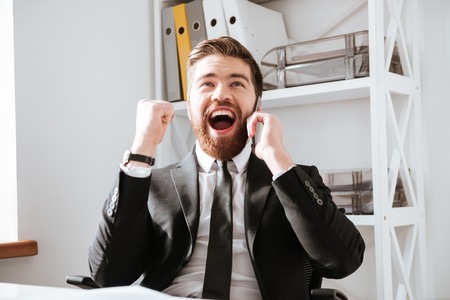 Image of happy businessman sitting in office while talking by phone and make winner gesture.の写真素材