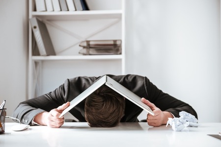 Image of young tired businessman lies on table in office with laptop computer on his head.の写真素材