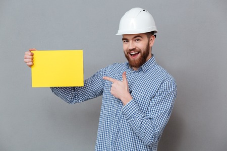 Happy man in helmet and shirt holding blank board and pointing at him in studio. Isolated gray backgroundの写真素材