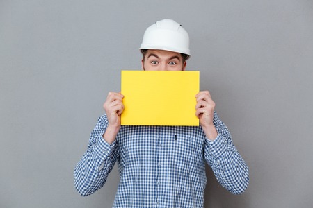 Man in helmet and shirt hiding behind the blank board in studio. Isolated gray backgroundの写真素材