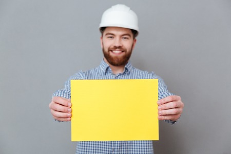 Photo of happy bearded man builder showing copyspace blank to camera standing over grey background. Focus on blank.の写真素材