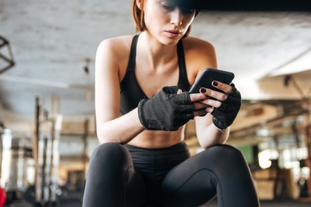 Beautiful young woman athlete sitting and using smartphone in gymの写真素材
