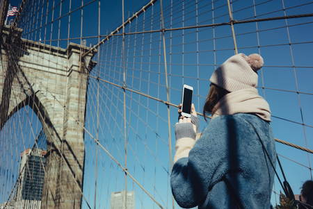 Side view of a tourist woman taking picture with smart phone while standing on Brooklyn Brige, New York City, Manhattan, USAの写真素材