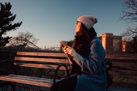 Smiling young woman in coat holding mobile phone while sitting on a bench and looking awayの写真素材