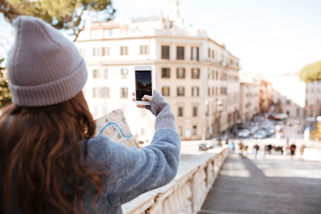 Back view of woman tourist taking pictures with blank screen smartphone in the cityの写真素材