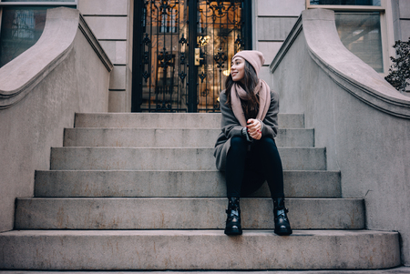 Full length portrait of a smiling woman in hat sitting on the house stairs outdoors and looking awayの写真素材