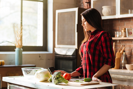 Young Woman in red shirt standing in kitchen with tablet computer and looking recipes. Side viewの写真素材