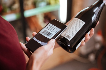 Closeup of woman scanning bar code with mobile phone on wine bottle in grocery storeの写真素材