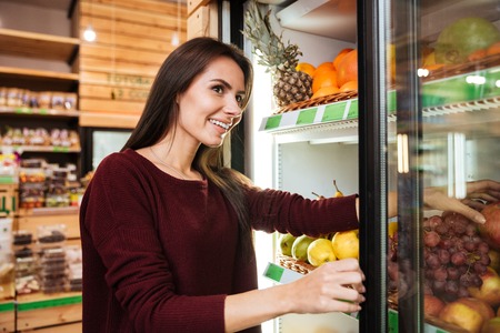 Smiling pretty young woman choosing and buying fruits at grocery shopの写真素材