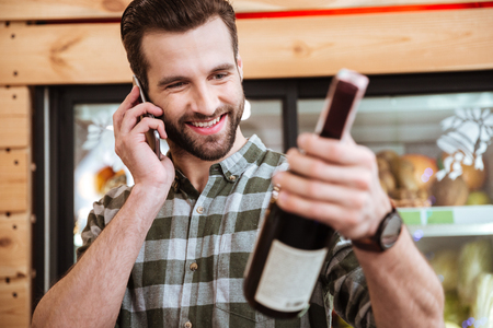 Happy young man buying wine and talking on cell phone in grocery shopの写真素材