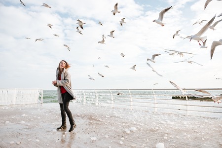 Happy pretty young woman standing and looking at seagulls in winterの写真素材